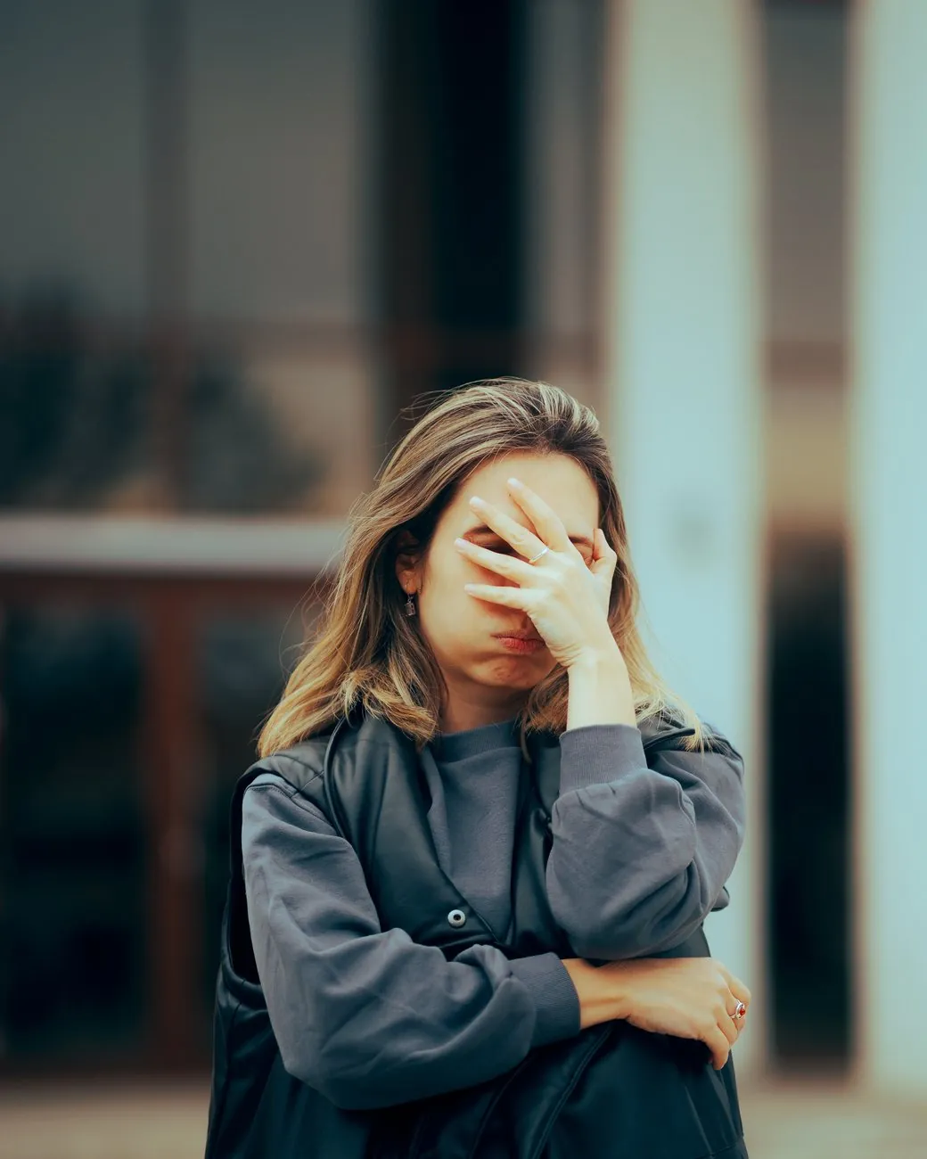 A woman stands outside a building with her eyes closed and one hand pressed to her face, her other arm folded across her body.