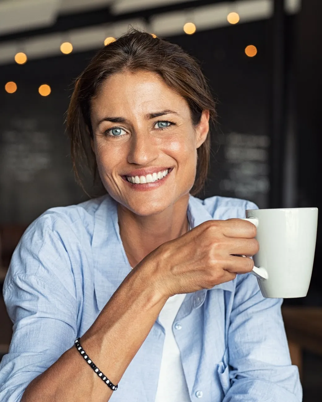 A woman sits in a softly lit cafe, smiling warmly at the camera with a white mug in her hand.